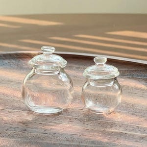 May include: Two miniature clear glass jars with matching removable lids. Each jar features a rounded body, a short neck, and a small knob handle on the centre of the lid. The jars are transparent and smooth, showcasing a simple, functional design.