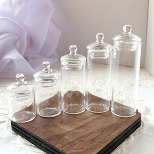 May include: Five clear glass jars with rounded lids of varying heights are arranged on a dark wooden base. The jars are empty and transparent, allowing a view of the background. The lids have a small, spherical knob on top.