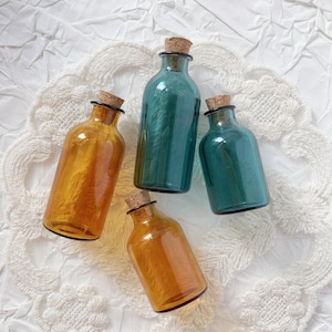 May include: Four decorative glass bottles in shades of amber and teal, each with a cork stopper. The bottles are arranged on a white, textured surface, creating a visually appealing display. These bottles are ideal for home decor.