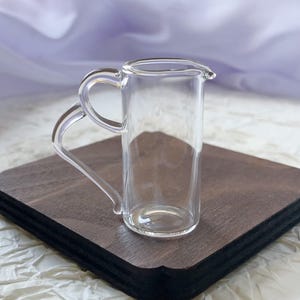 Op de afbeelding: Een heldere glazen roomkannetje met een handvat en een tuit, geplaatst op een donkerbruin houten dienblad. Het kannetje is leeg en lijkt van dun glas te zijn. De achtergrond is een zachte, vage stof.