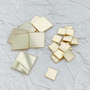 May include: A collection of small, square, gold-colored metal tiles scattered on a white, textured surface. The tiles are reflective and appear to be made of a shiny material, possibly for crafting or decorative purposes.