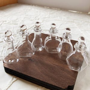 Puede incluir: Colección de seis botellas de vidrio transparente con tapones redondeados. Las botellas se exhiben en una bandeja de madera marrón oscuro. Las botellas tienen diferentes formas, incluyendo diseños redondos, de lágrima y hexagonales.