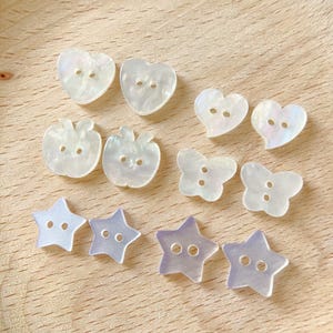 Puede incluir: Una colección de botones iridiscentes en varias formas: corazones, manzanas, mariposas y estrellas. Cada botón tiene dos agujeros para coser. Los botones están dispuestos sobre una superficie de madera clara.