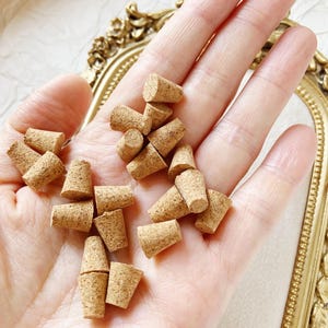 Könnte beinhalten: Eine Ansammlung kleiner, konisch zulaufender Korken, wahrscheinlich für Flaschen, wird in einer Hand gehalten. Die Korken sind hellbraun und haben eine strukturierte Oberfläche. Im Hintergrund befindet sich ein goldfarbener, verzierter Rahmen.