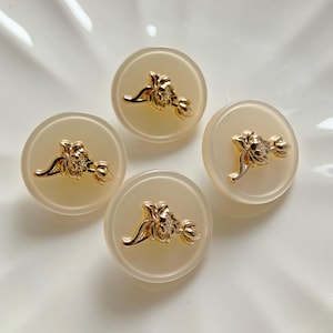 以下が含まれることがあります： 金色の花の模様が付いた、丸いオフホワイトのボタンが4つ。ボタンはわずかに半透明で、花の装飾が施されています。白い表面に配置されています。