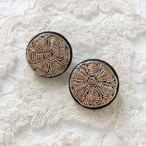 May include: Two round, gold-colored metal earrings with a floral design. The earrings are set on a black fabric base. The background is a white, lacy fabric.