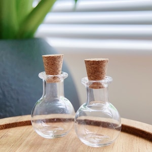 Puede incluir: Dos pequeñas botellas de vidrio transparente con tapones de corcho. Las botellas tienen forma redondeada y están sobre una superficie de madera. El fondo incluye una planta verde y una persiana de lamas blancas.