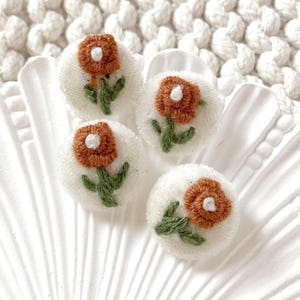 Può includere: Quattro piccole toppe rotonde di tessuto bianco con fiori ricamati. Ogni fiore ha un motivo a petali color ruggine, un centro bianco e foglie e steli verdi. Le toppe sono disposte su una superficie bianca smerlata.
