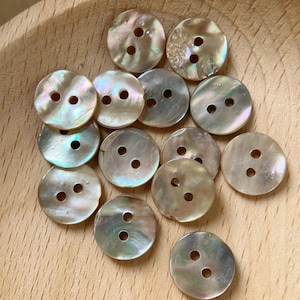 May include: A selection of iridescent, circular buttons, each with two holes. The buttons display a pearlescent sheen, reflecting hues of green, pink, and gold. They are arranged on a light brown wooden surface.