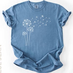 May include: A blue jean colored short-sleeve t-shirt featuring a white graphic print of three dandelion flowers with seeds blowing in the wind across the chest. The shirt has a crew neckline and rolled sleeves, displaying a casual, textured fabric.