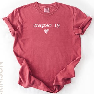 May include: Crimson-colored t-shirt with rolled-up sleeves. The shirt features the text "Chapter 19" above a small heart graphic in white. The word "CRIMSON" is printed vertically on the left side of the shirt.