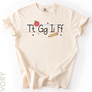 May include: Ivory-colored t-shirt featuring a school-themed design. The design includes the letters Tt Gg Ii Ff, a red apple, a yellow pencil, and small black stars. The shirt is made of a soft material.
