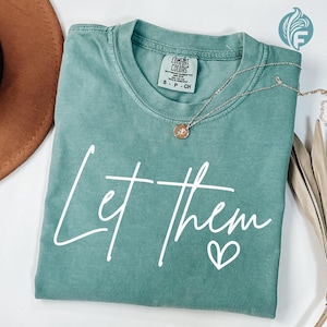 May include: A teal blue t-shirt with the text "Let them" in white script with a small heart underneath. The shirt is made of a soft, comfortable material.
