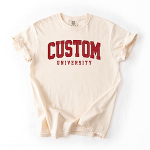 May include: Cream-colored t-shirt with the word "CUSTOM" in bold, red, collegiate-style lettering above the word "UNIVERSITY" in a smaller, matching font. The shirt has rolled-up sleeves and a classic crew neck.