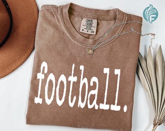 May include: A brown Comfort Colors t-shirt with the word "football" printed in white. The shirt has a crew neck and short sleeves. A silver necklace with a small pendant is draped across the shirt. A brown hat is visible in the upper left corner.
