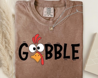 May include: A brown Comfort Colors t-shirt with the word "GOBBLE" in black, featuring a cartoon turkey face. A silver necklace with a pendant is draped across the shirt. The shirt has a crew neck and short sleeves. The shirt is on a neutral background.