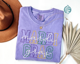 May include: A light purple t-shirt with the words "Mardi Gras" in white script and outlined in green, yellow, and purple. A gold necklace and a brown hat are also in the image.