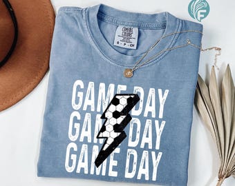 May include: A light blue t-shirt with the words "GAME DAY" in white, repeated three times. A black and white soccer ball design is in the shape of a lightning bolt. A gold necklace with a pendant is on the shirt.