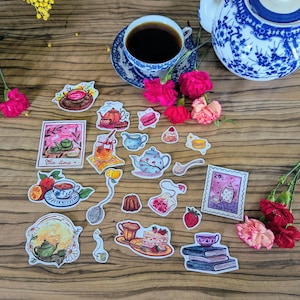 Peut inclure: Divers autocollants sur le thème du thé et une tasse de thé sur une surface en bois. Les autocollants représentent des théières, des tasses à thé, des pâtisseries et le texte "Tea Time". Des fleurs roses et une théière bleue et blanche sont également présentes.