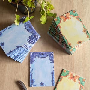 Peut inclure: Plusieurs blocs-notes décoratifs avec des motifs de papillons et de fleurs en bleu, jaune et vert. Les blocs-notes rectangulaires présentent des bordures ornées. Un stylo argenté repose sur la surface en bois près des blocs-notes.