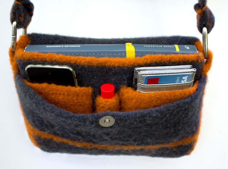 Pu&ograve; includere: Una borsa a tracolla in maglia a righe grigie e arancioni con chiusura a zip e bottone a pressione. La borsa &egrave; aperta e ha diverse tasche all'interno. Le tasche contengono un telefono, un taccuino, un rossetto e carte di credito.