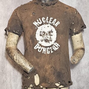 May include: Brown distressed t-shirt featuring the white text "NUCLEAR DUNGEON" encircling a white gas mask and skull graphic. The shirt has a vintage appearance with holes and frayed edges, giving it a worn look.