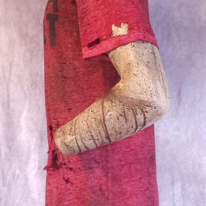 Pu&ograve; includere: Una maglietta rossa usurata con la lettera "T" visibile. La maglietta presenta fori e un aspetto usurato. Il braccio del manichino &egrave; visibile, con un aspetto strutturato e invecchiato. L'estetica generale suggerisce uno stile vintage o grunge.