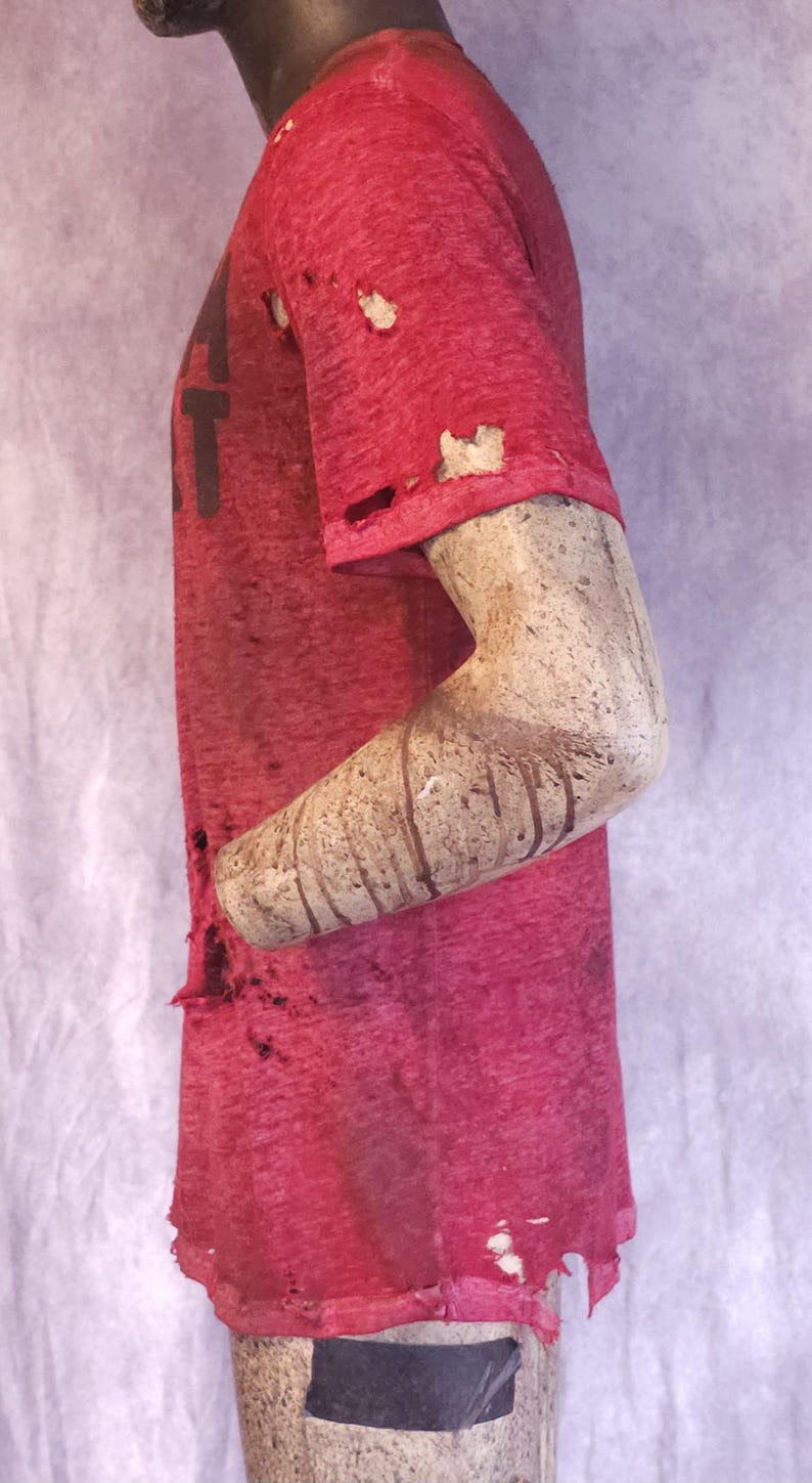 Pu&ograve; includere: Una maglietta rossa usurata con la lettera "T" visibile. La maglietta presenta fori e un aspetto usurato. Il braccio del manichino &egrave; visibile, con un aspetto strutturato e invecchiato. L'estetica generale suggerisce uno stile vintage o grunge.