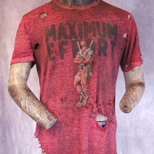 Pu&ograve; includere: T-shirt rosso con la scritta "MAXIMUM EFFORT" e una grafica di Deadpool. La maglietta ha un aspetto invecchiato con fori e bordi sfilacciati, che le conferiscono un aspetto vintage.