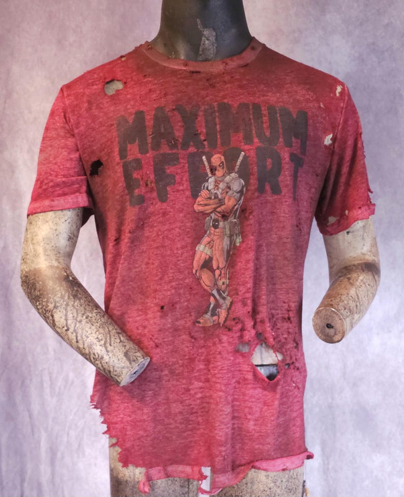 Pu&ograve; includere: T-shirt rosso con la scritta "MAXIMUM EFFORT" e una grafica di Deadpool. La maglietta ha un aspetto invecchiato con fori e bordi sfilacciati, che le conferiscono un aspetto vintage.