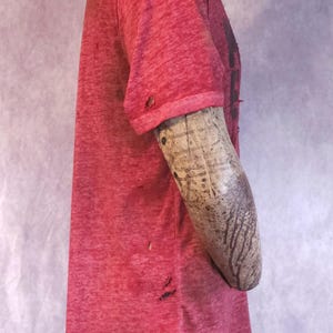 Pu&ograve; includere: Una maglietta rossa consumata con girocollo e maniche corte. La maglietta ha un aspetto usurato e strutturato con diversi piccoli fori e un orlo sfilacciato. La maglietta &egrave; esposta su un manichino.