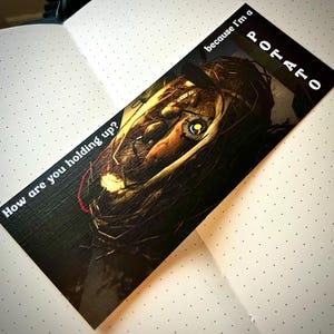 May include: A bookmark with a dark, textured background featuring a close-up of a doll's face. The text "How are you holding up?" is on the left side of the bookmark and "because I'm a POTATO" is on the right side.