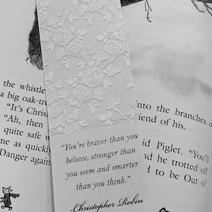 Puede incluir: Un marcapáginas blanco con un diseño floral y una cita de Christopher Robin: "Eres más valiente de lo que crees, más fuerte de lo que pareces y más inteligente de lo que piensas."