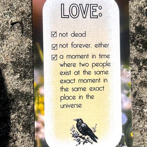 May include: A rectangular bookmark with the word "LOVE:" at the top. The text includes "not dead," "not forever," and a definition of love. A black bird illustration is at the bottom, with "The Dead Romantics" text.