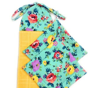 May include: A kitchen set featuring a yellow hand towel and matching pot holders. The pot holders and the top of the towel have a teal floral pattern with red, purple, and yellow flowers. The towel has a loop for hanging.