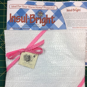 May include: A white square piece of Insul-Bright needled insulated lining with a pink polka dot ribbon tied around it. The ribbon is tied in a bow with a small tag that says "She Who Sews 698". The product is labeled "Insul-Bright" and "Conserve Body Warmth".