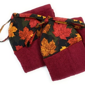 May include: Two burgundy hand towels with a decorative top featuring a fall leaf pattern in red, orange, and yellow on a black background. The towels are tied together with a matching fabric loop.