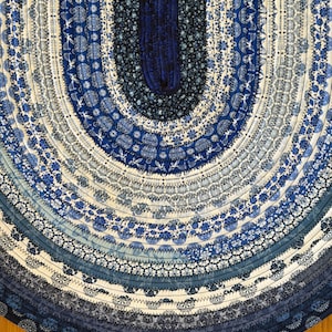 May include: An oval-shaped rug constructed from coiled fabric strips in various shades of blue and white. The textile features a repeating pattern of floral, geometric, and bird motifs stitched together with visible dark thread in a concentric design.