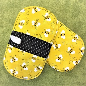 May include: A pair of yellow oven mitts with a black and white polka dot trim. The mitts are decorated with a repeating pattern of bees and have a white loop for hanging.