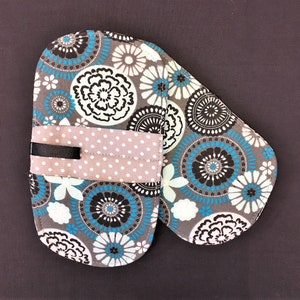 May include: A gray oven mitt with a floral pattern in white, teal, and black. A light pink band with white polka dots and a black ribbon detail is sewn across the mitt.