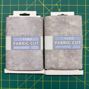 May include: Two folded rectangular packages of gray fabric, each labeled 1 Yard Fabric Cut and 100% Cotton. The labels indicate dimensions of 36x44 inches or 91.4x111.8 cm. The fabric has a mottled, textured appearance in shades of light and dark gray.