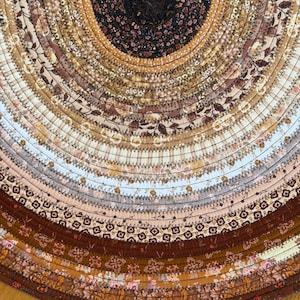 May include: A circular rug constructed from coiled fabric strips featuring a variety of floral and geometric patterns in a color palette of black, dark brown, tan, cream, and white. The fabric coils are joined together with dark zigzag stitching.