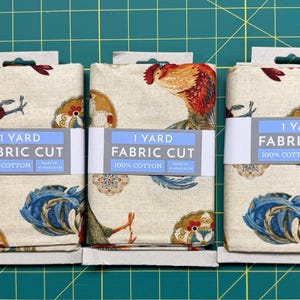 May include: Three folded fabric cuts, each labeled 1 YARD FABRIC CUT 100% COTTON. The beige fabric features a repeating pattern of roosters with red, orange, and blue feathers, interspersed with circular floral motifs in shades of gold and blue.
