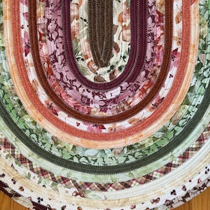 May include: An oval-shaped braided fabric rug featuring concentric layers of cotton strips in various autumn-inspired patterns. The design includes shades of deep burgundy, forest green, burnt orange, cream, and brown with leaf and plaid motifs.