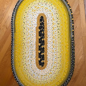 May include: An oval-shaped braided rug featuring concentric rings of fabric in shades of yellow, white, and black. The pattern includes floral prints, honeycomb motifs, and a black and white gingham border. The center is a solid black fabric.