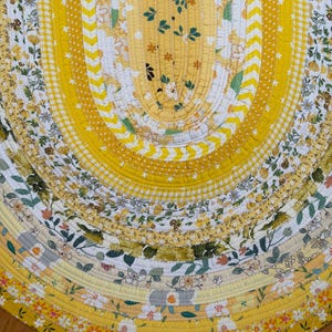 May include: An oval-shaped rug constructed from coiled fabric strips in various shades of yellow, white, and green. The design features alternating bands of floral patterns, chevron stripes, and gingham checks, all stitched together with visible thread.