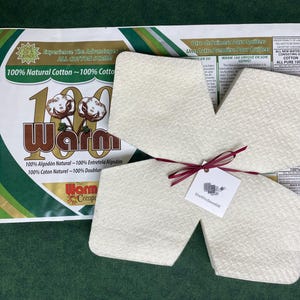 May include: Off-white, textured cotton warmers tied with a burgundy ribbon, with a tag attached. A package with text reading "100% Natural Cotton" and "Warm" is in the background. The background is a dark green.