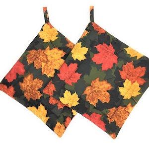 May include: Two square pot holders with a fall leaf pattern. The leaves are in shades of red, orange, and yellow against a dark green and black background. Each pot holder has a small fabric loop for hanging.