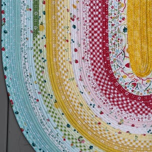 May include: A round fabric rug constructed from coiled strips of multi-colored cotton material. The design features concentric bands of various patterns including red gingham, yellow checks, and floral prints with strawberries and lemons.