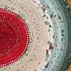 May include: An oval fabric mat constructed from coiled strips of cotton featuring a variety of holiday-themed patterns. The center is deep red with white speckles and gingham, transitioning outward through beige, light green, and dark green bands.